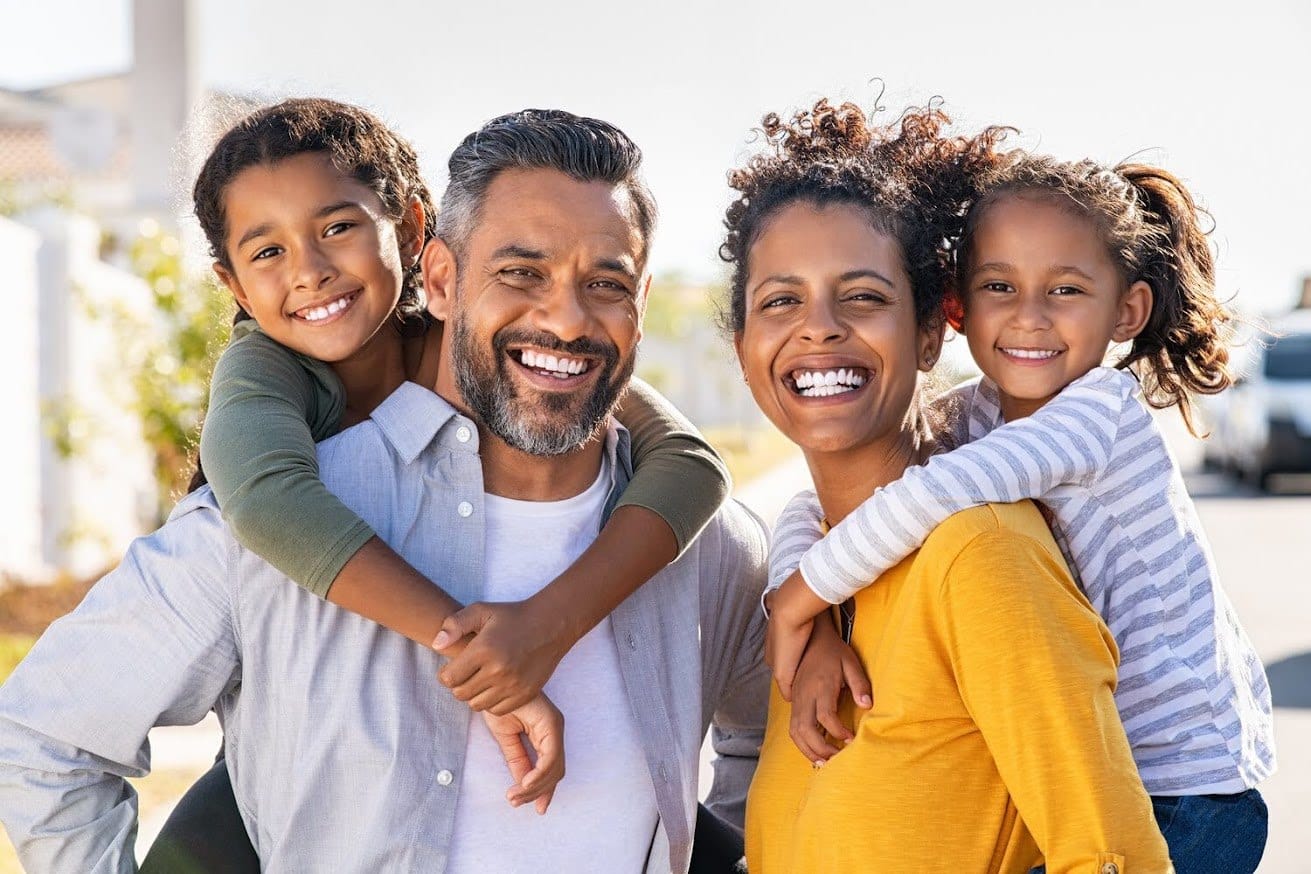 A smiling family of four, the children riding piggyback on their parents