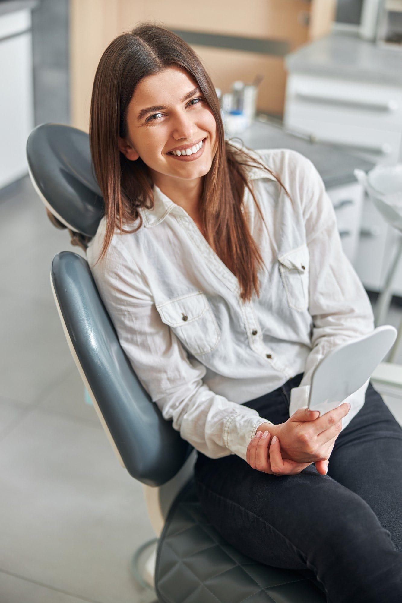 A smiling woman seated in a dental chair, holding a hand mirror