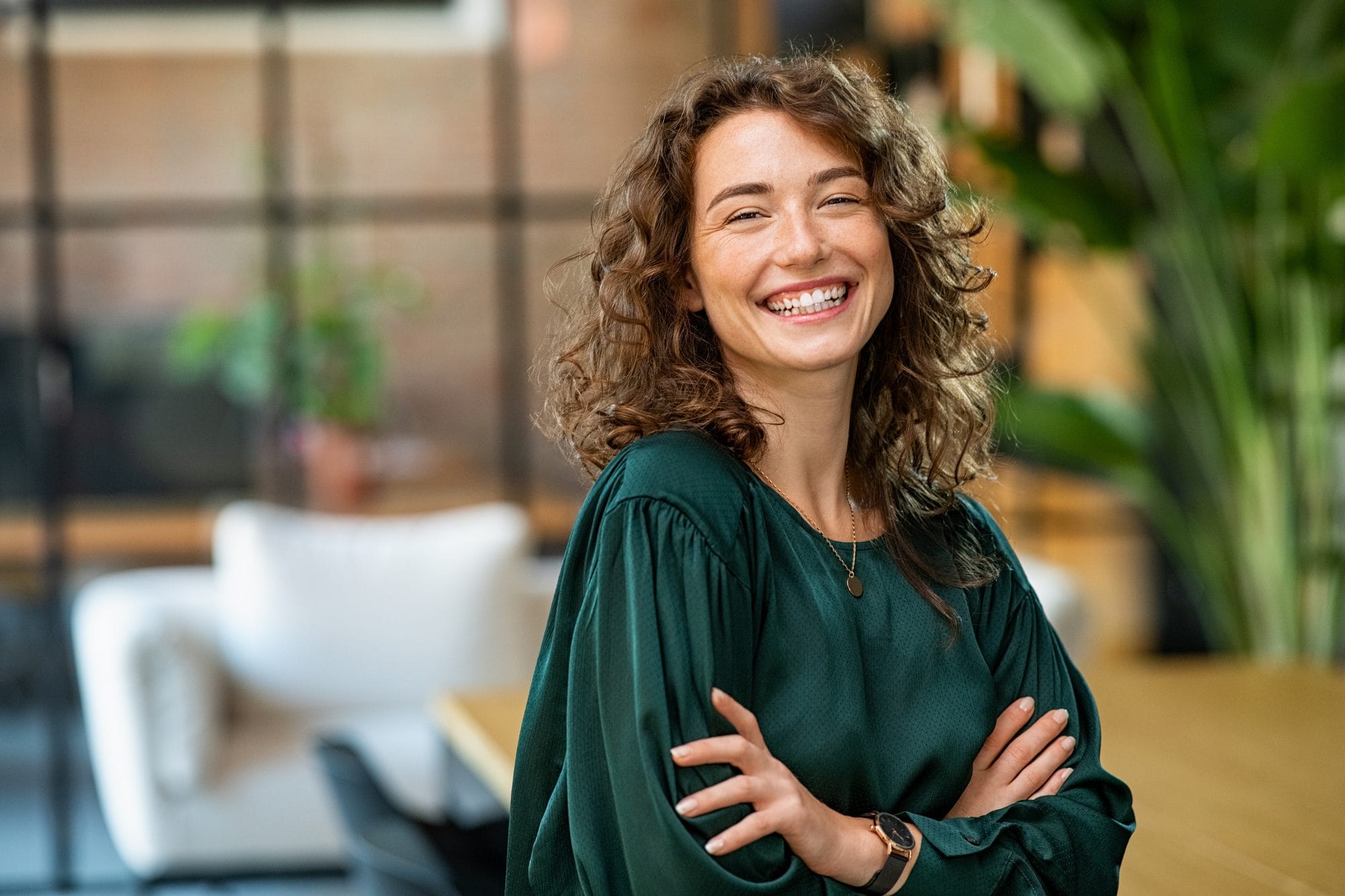 A smiling woman with curly brown hair and folded arms in a bright, plant-filled room